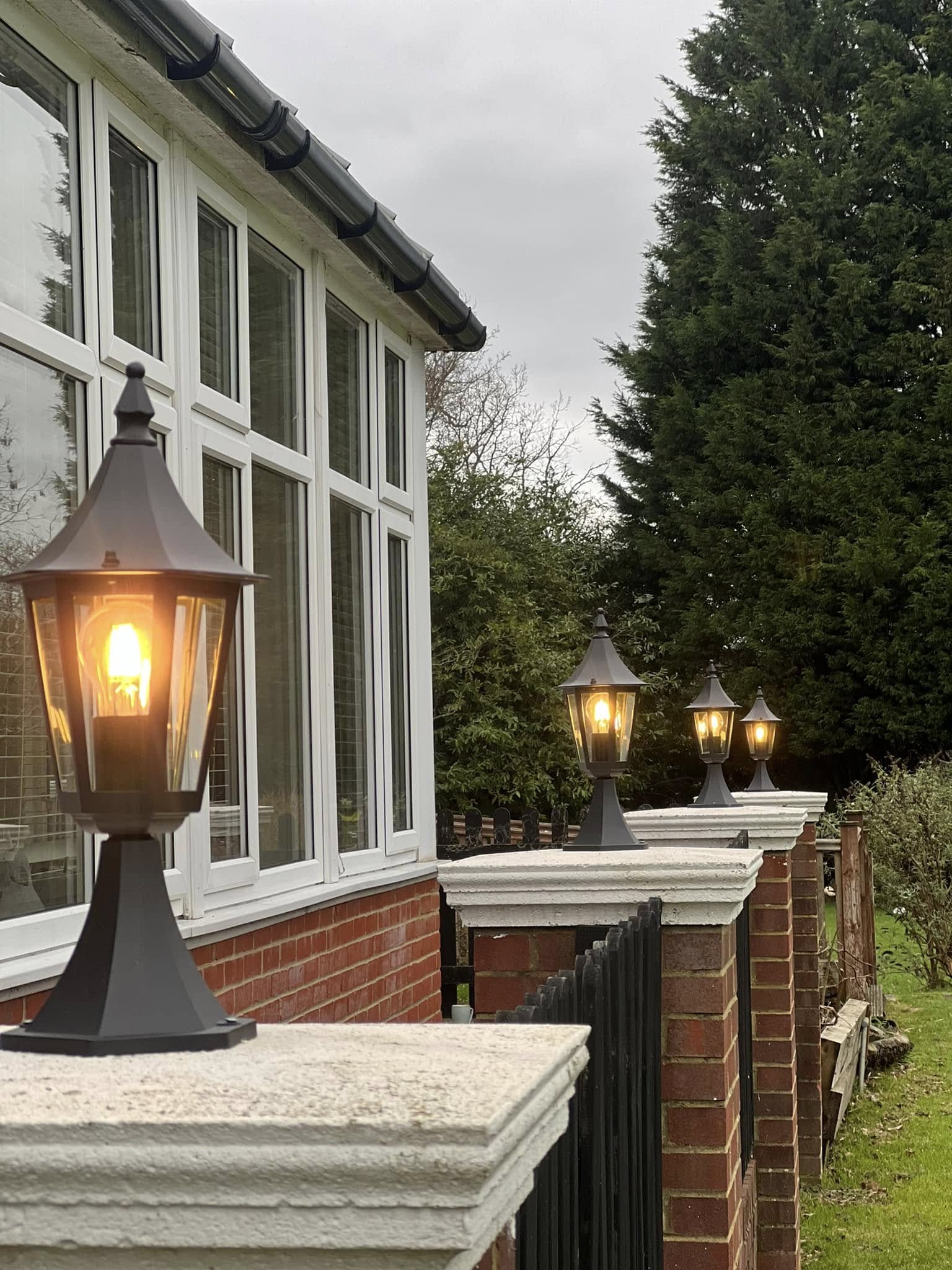 Four black lantern-style lights are mounted on brick pillars along a walkway beside a house with large windows and a tall tree in the background.