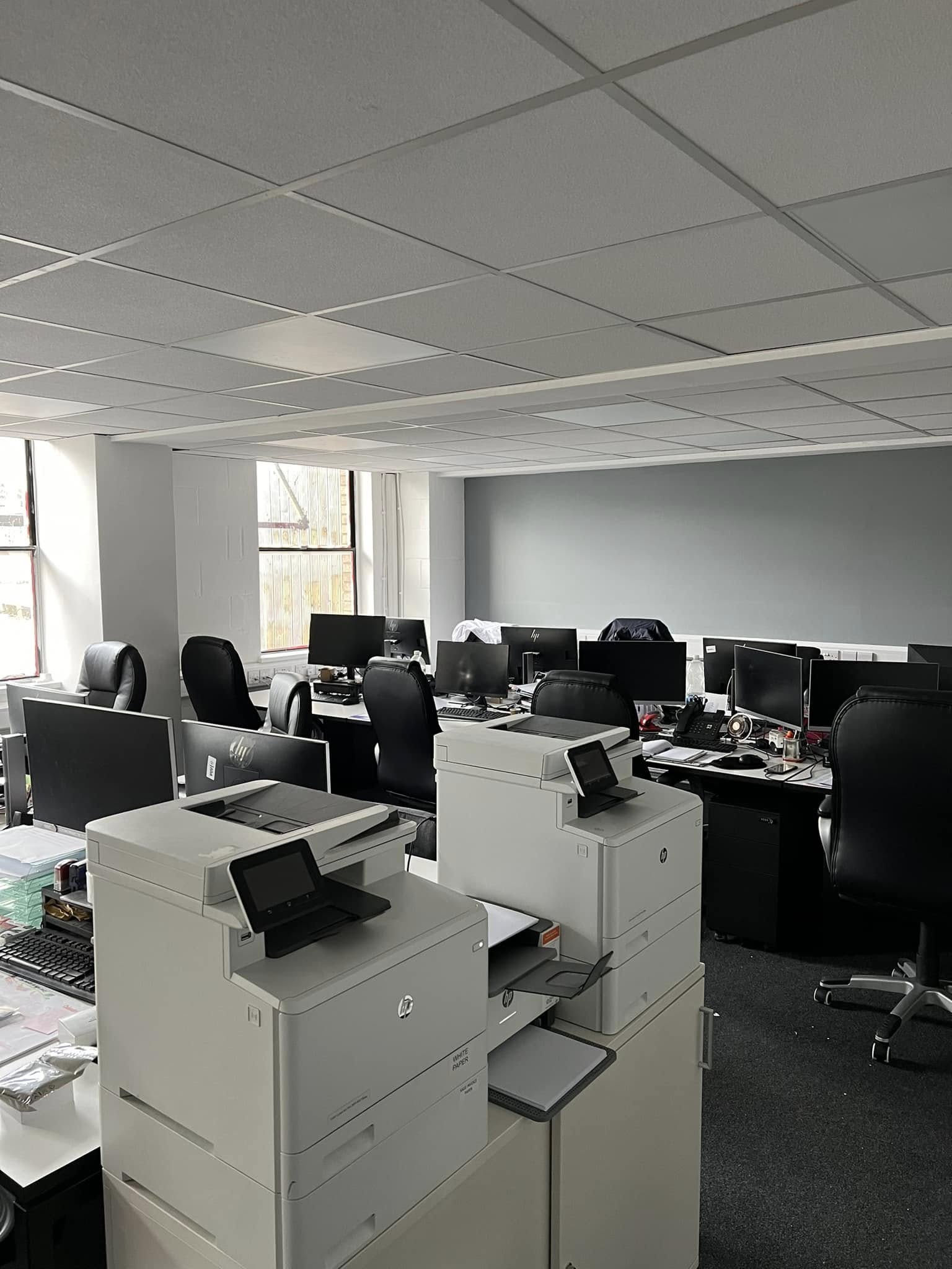 Modern office with several empty desks, black chairs, computer monitors, printers, and large windows letting in natural light.