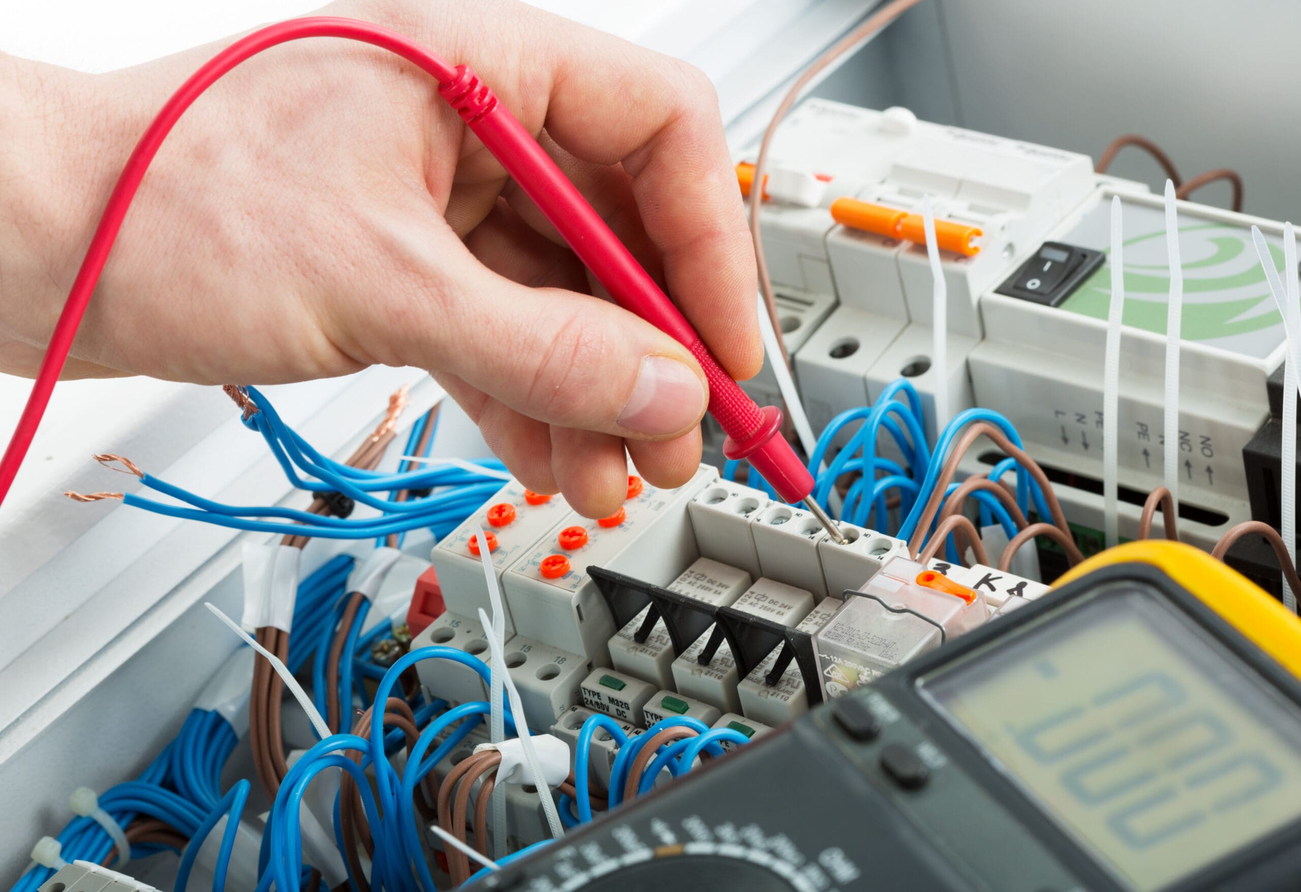 A hand uses a red test probe on an electrical panel with various wires and components; a digital multimeter is visible in the foreground.