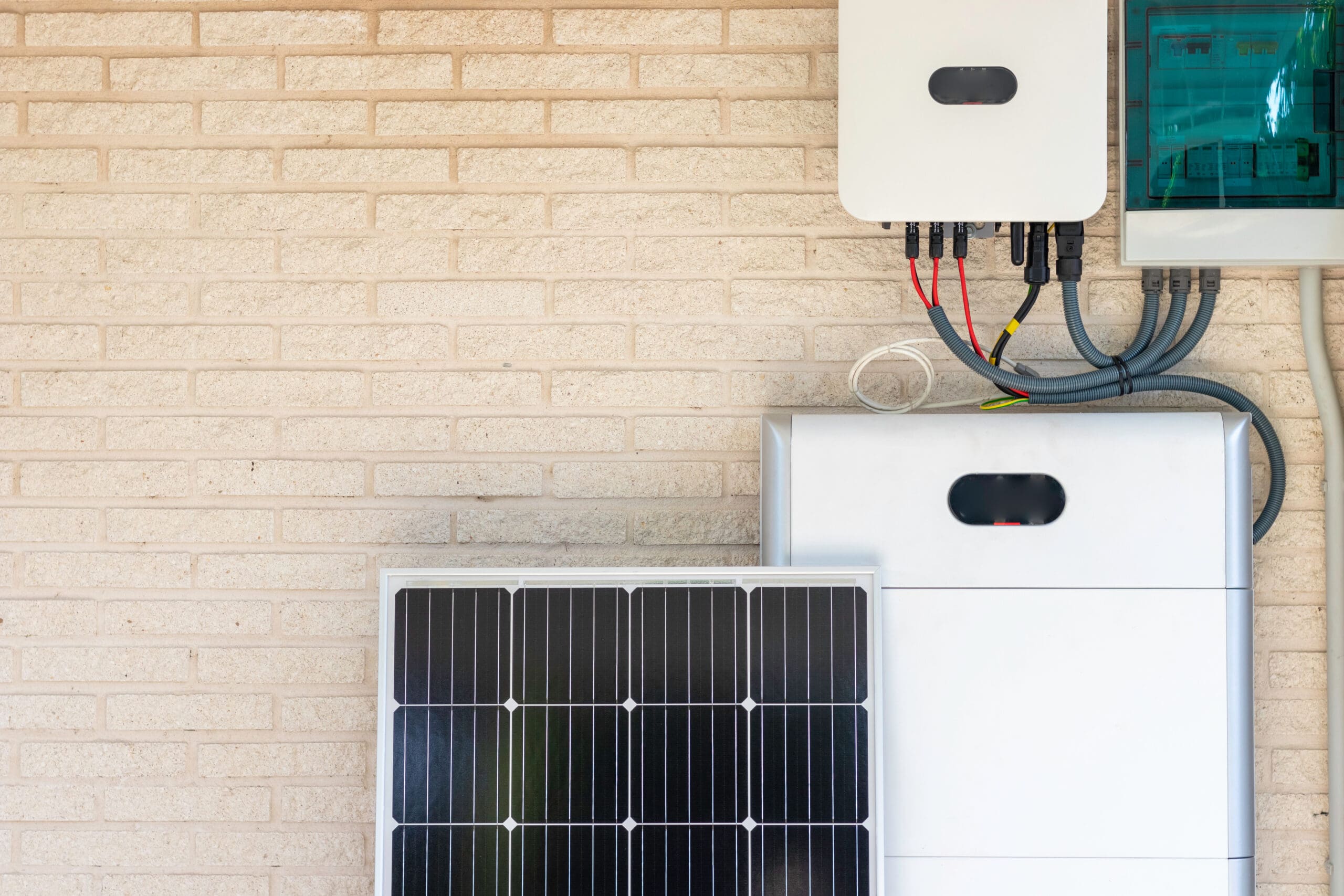 A solar panel leaning against a brick wall, next to an inverter, battery storage unit, and electrical consumer unit with visible wiring.