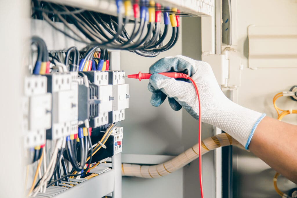 A gloved hand uses a red probe to test electrical connections inside an open electrical panel with multiple wires and components.
