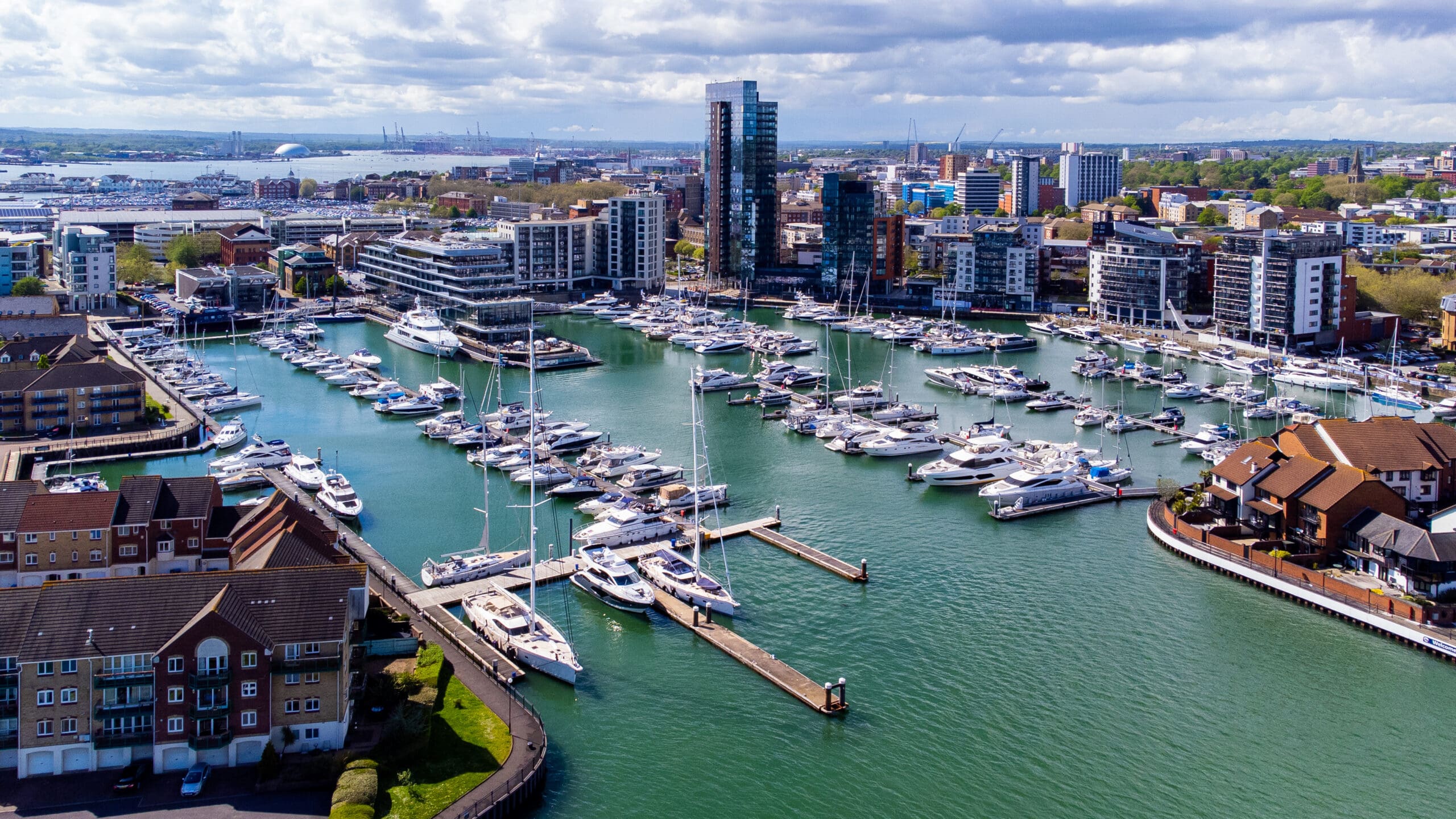 Aerial view of a marina with numerous moored boats and yachts, surrounded by modern apartment blocks and cityscape in the background.