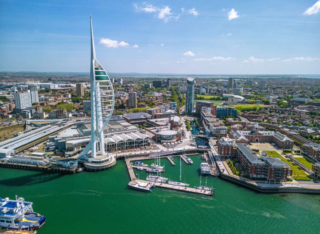 Aerial view of Portsmouth waterfront featuring the Spinnaker Tower, marina with moored boats, and surrounding city buildings on a clear day.