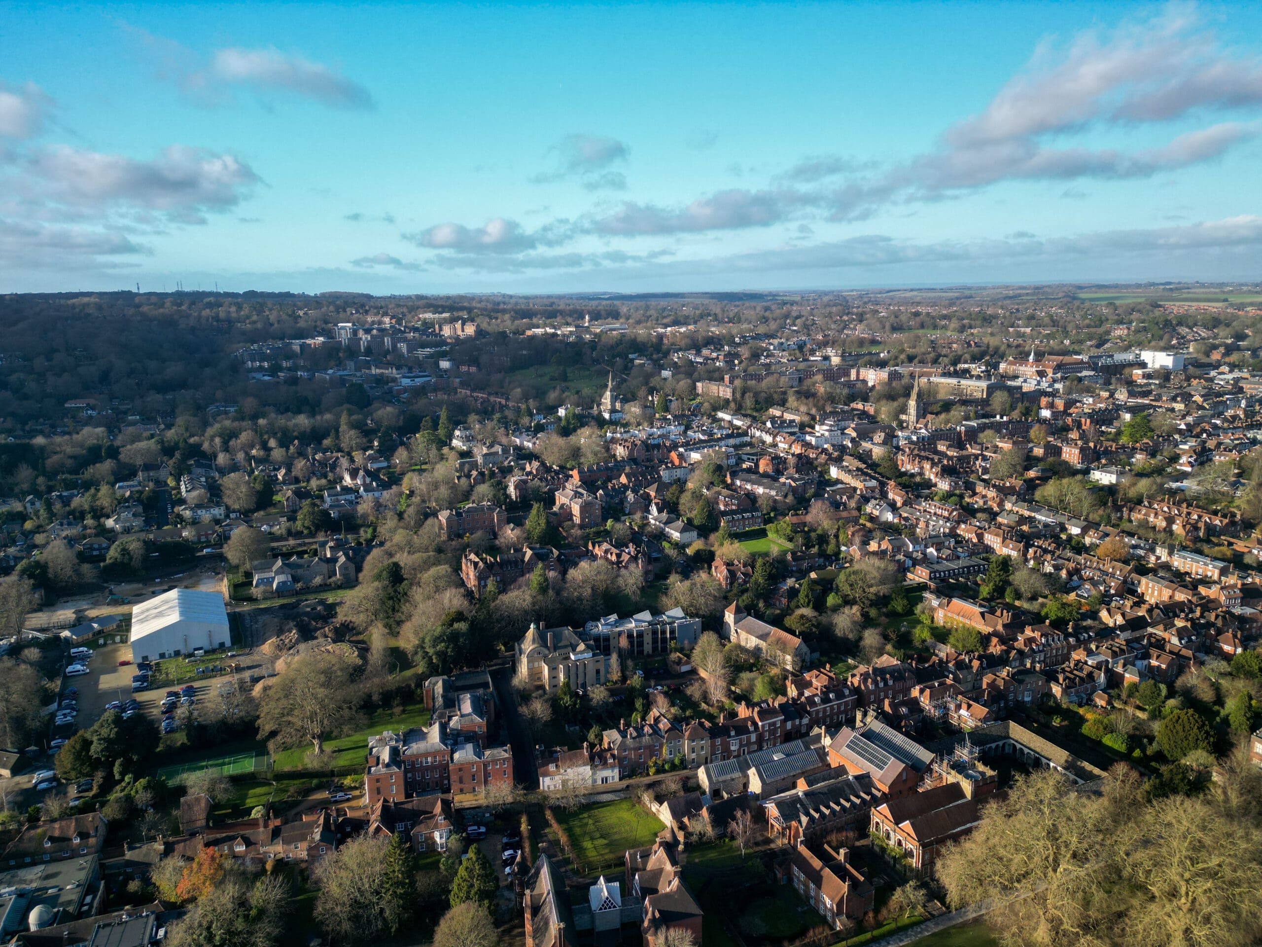 Aerial view of a suburban town with residential houses, trees, and scattered buildings under a partly cloudy sky.