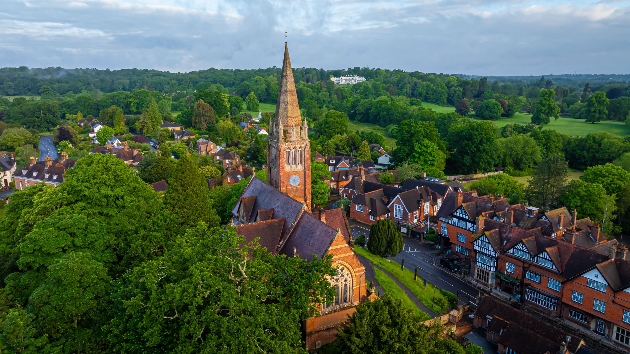 Aerial view of a brick church with a tall spire, surrounded by trees and traditional houses in a green, rural village setting.