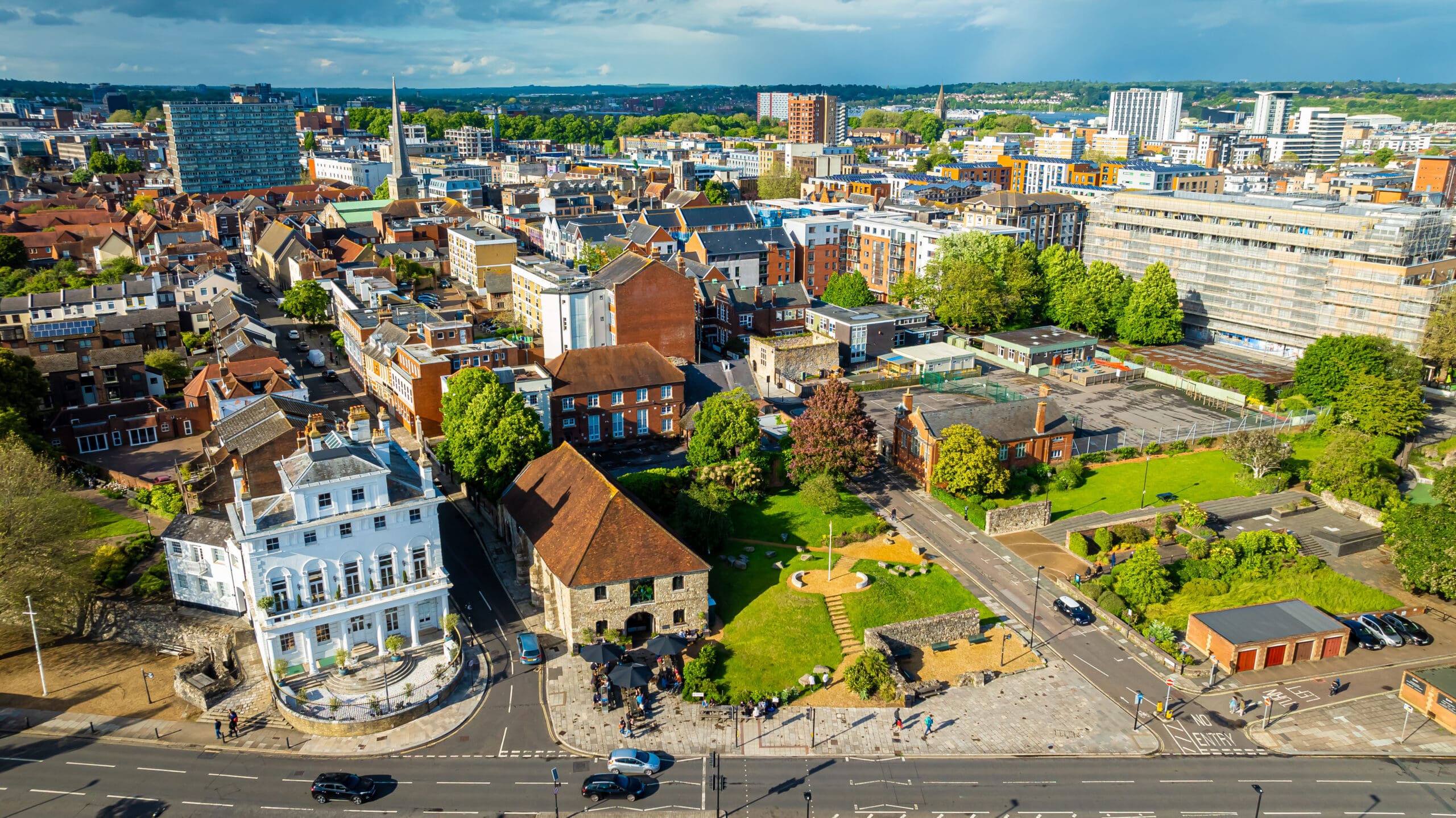 Aerial view of a cityscape with a mix of historic and modern buildings, green spaces, and people walking along streets and pathways on a sunny day.