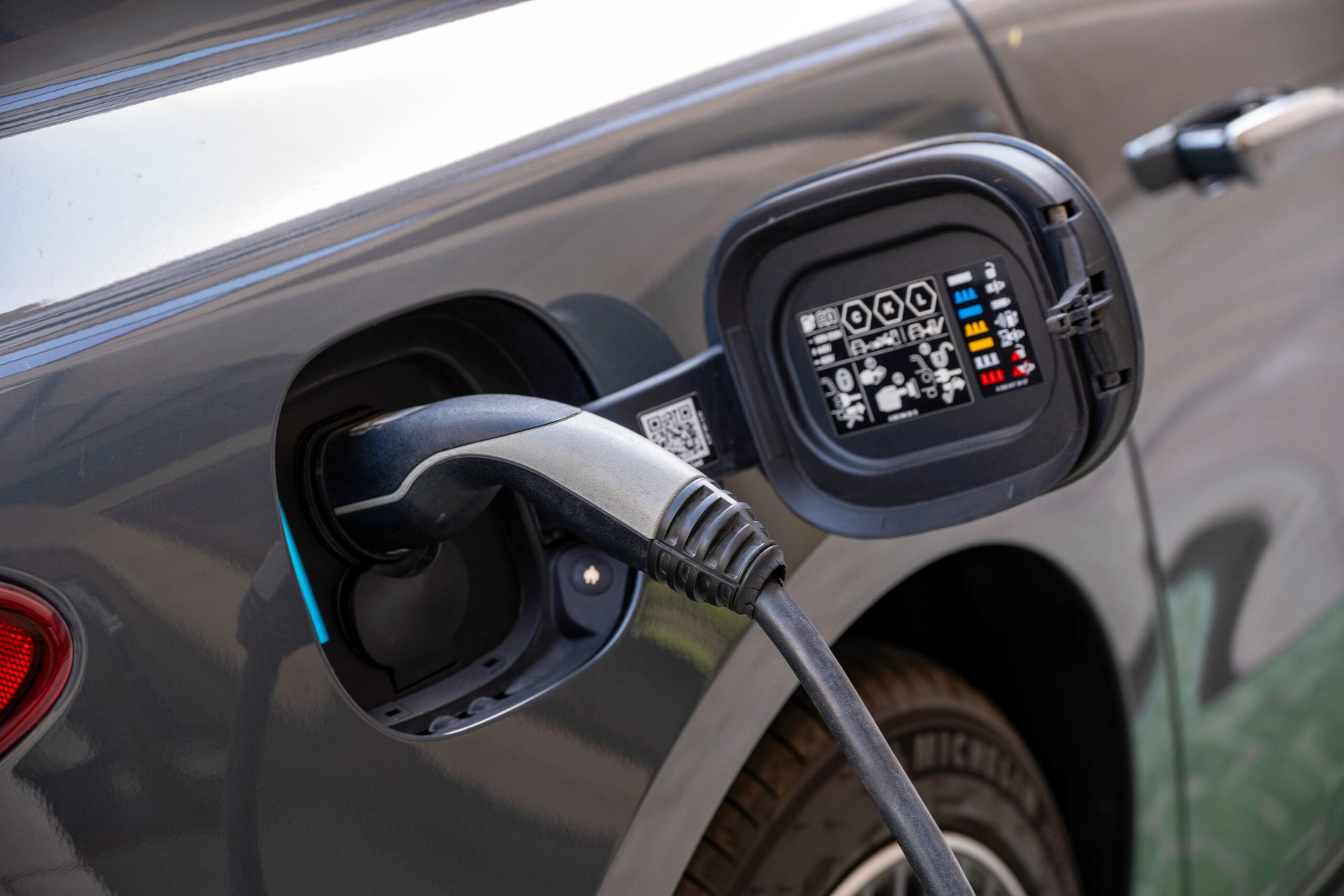 A black electric car is plugged into a charging station, with the charging port cover open and charging instructions visible on the inside.