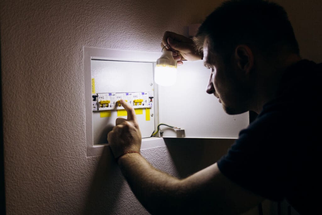 A person examines a fuse box in a wall, holding a small light bulb to illuminate the circuit breakers.