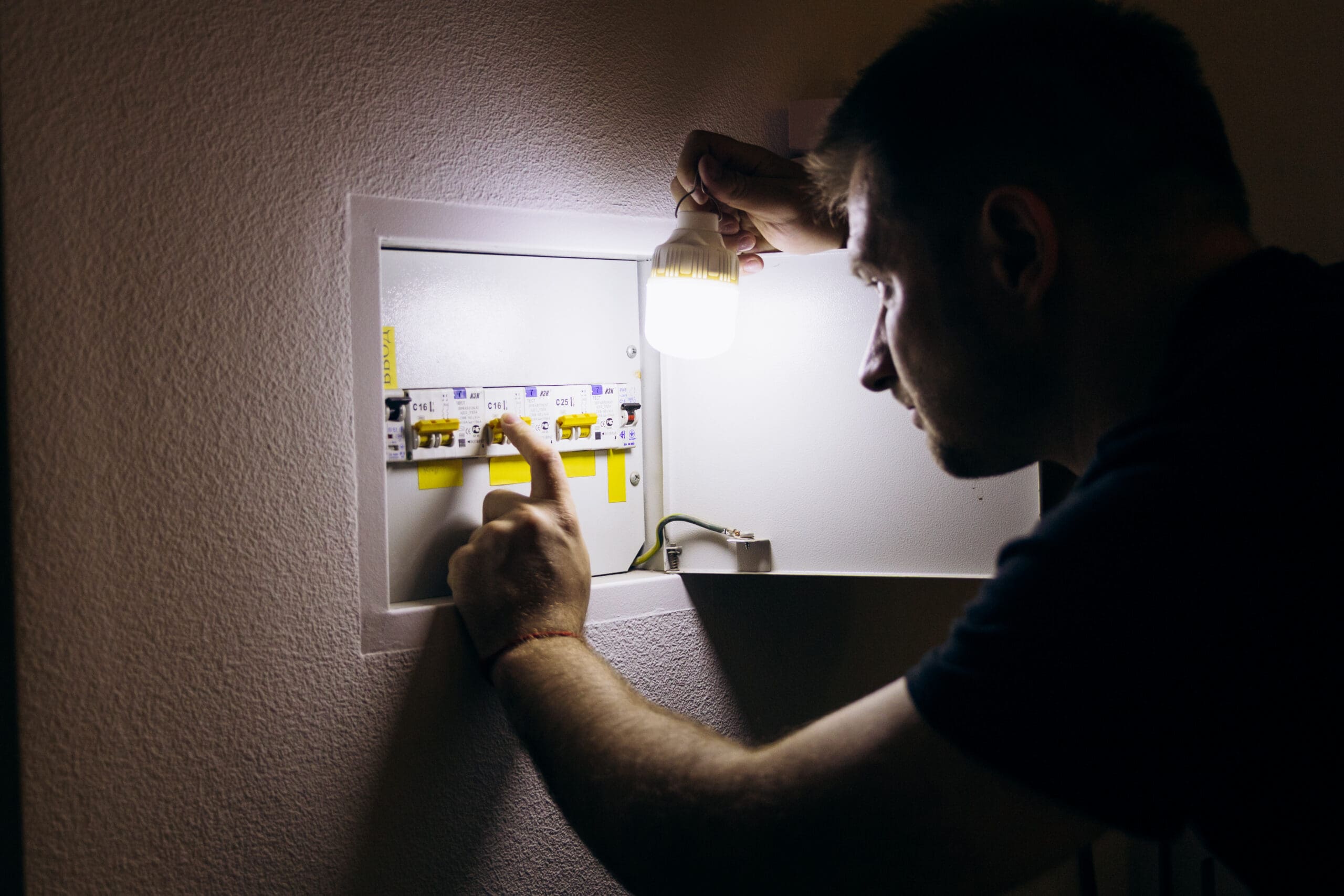 A person examines a fuse box in a wall, holding a small light bulb to illuminate the circuit breakers.
