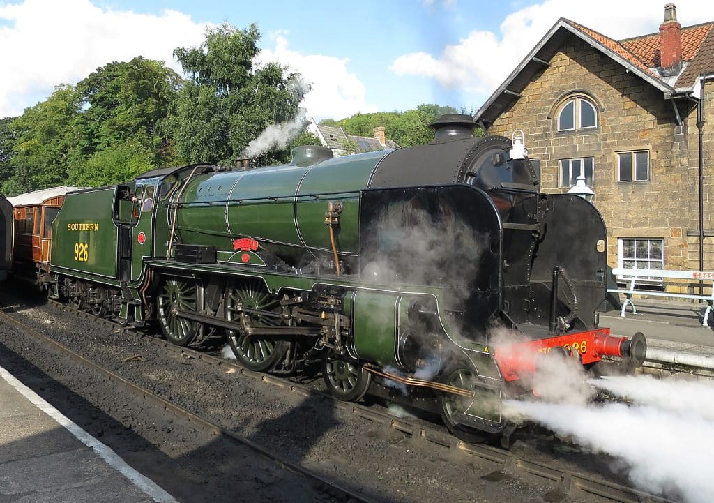 A green steam locomotive numbered 928 with “Southern” painted on the side, emitting steam at a railway station next to a brick building.